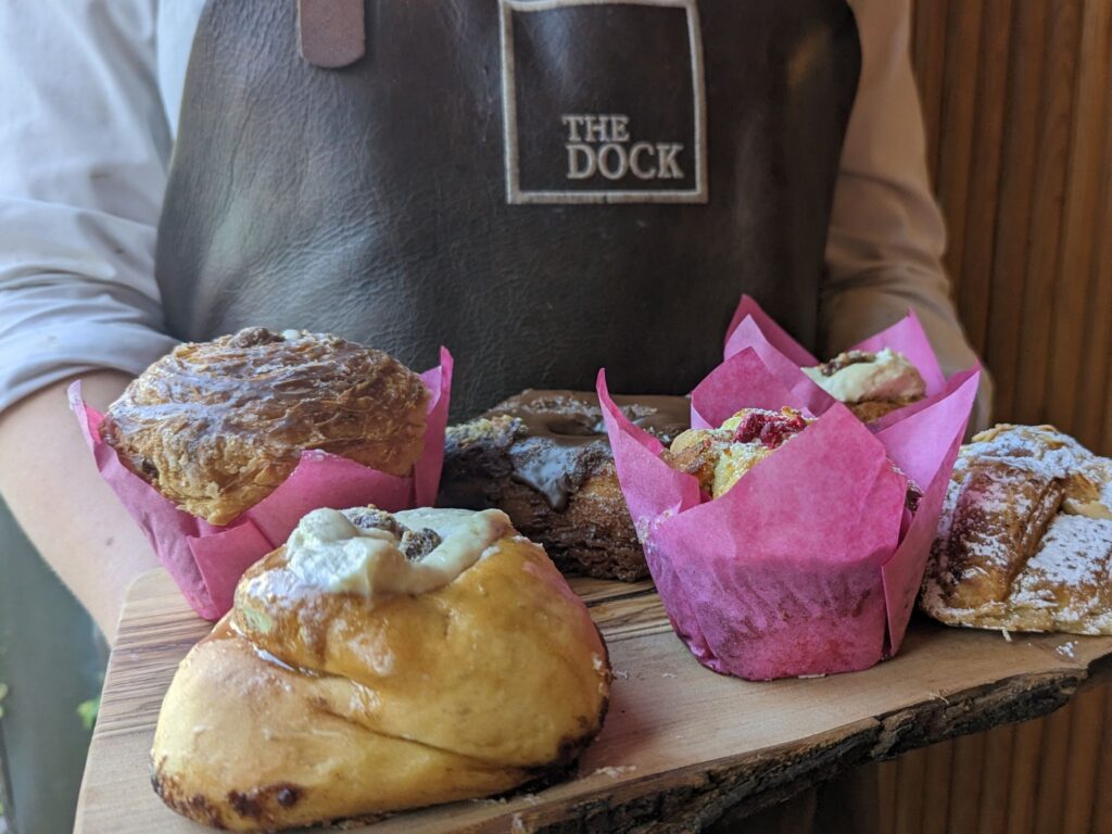 A person wearing a brown leather apron with “The Dock” logo holds a wooden serving board topped with assorted pastries, including muffins wrapped in pink paper, a glazed bun, a chocolate-covered pastry, and a sugar-dusted pastry.