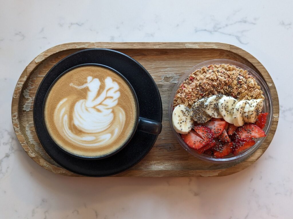 A wooden serving tray holds a black cup of latte with swan-shaped latte art beside a bowl of granola topped with sliced bananas, strawberries, and chia seeds. The tray rests on a light marble surface.
