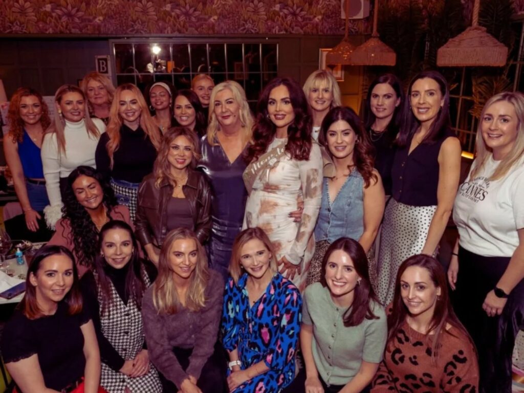 A group of women poses together indoors, all smiling warmly. They're dressed in a variety of stylish outfits, conveying a joyful and celebratory mood.