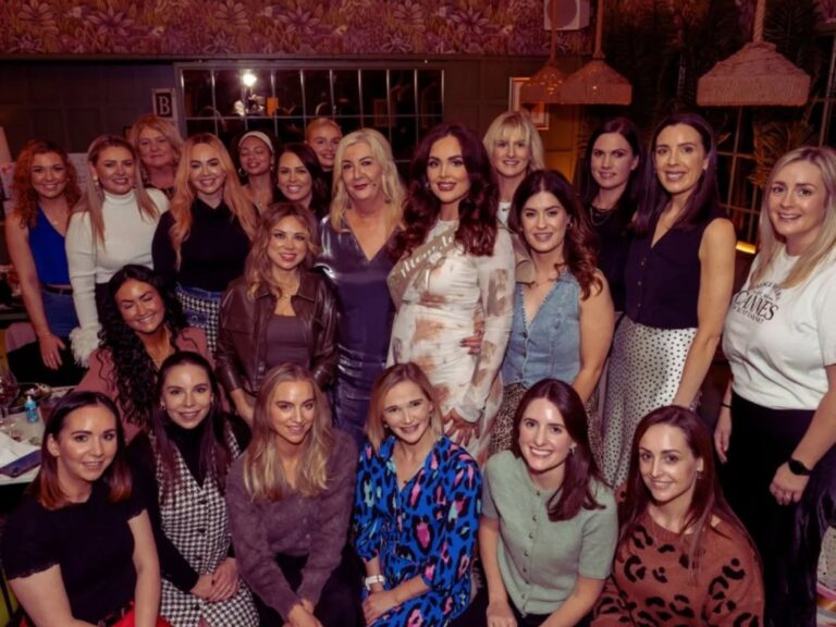 A group of women poses together indoors, all smiling warmly. They're dressed in a variety of stylish outfits, conveying a joyful and celebratory mood.