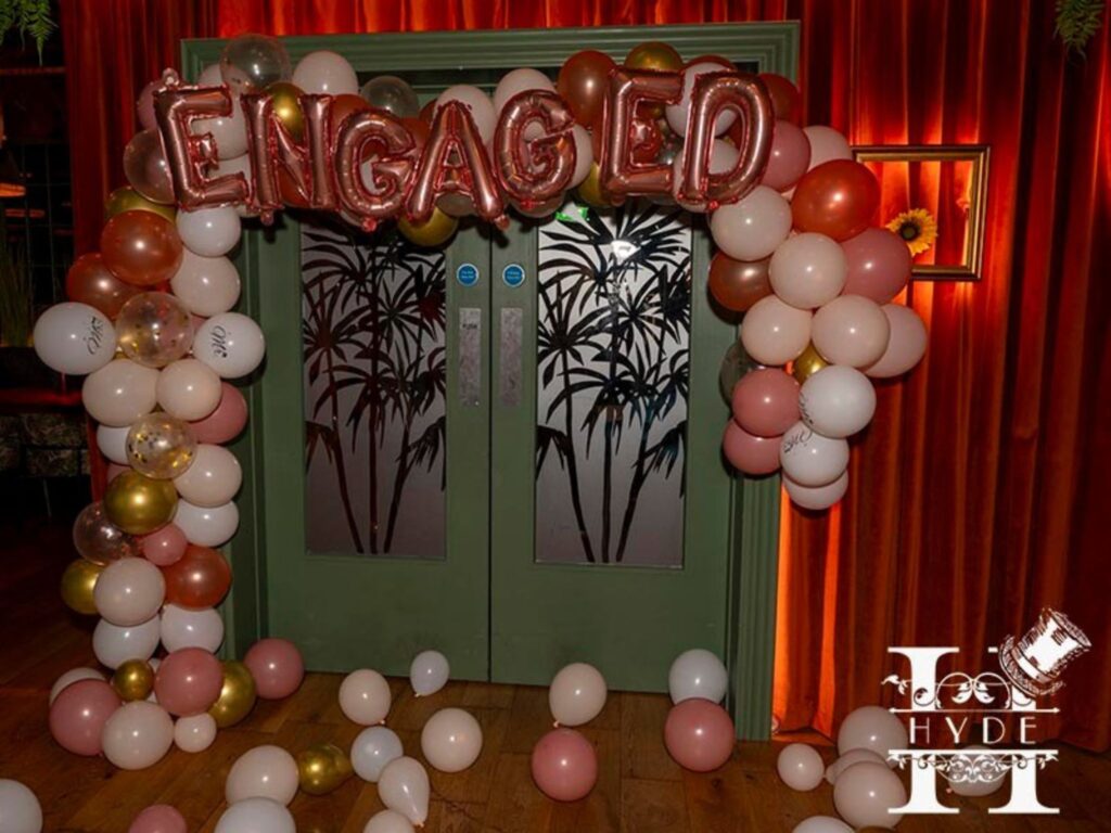 Engagement celebration entrance with pink, white, and gold balloon arch spelling "ENGAGED." Red curtains and decorated doors create a festive atmosphere.