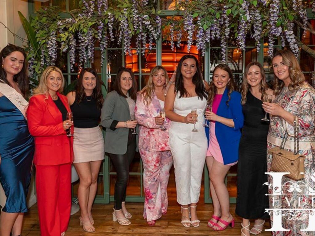 A group of nine women dressed in colorful outfits, smiling and holding glasses, stand under purple flowers in a lively indoor setting with festive decor.
