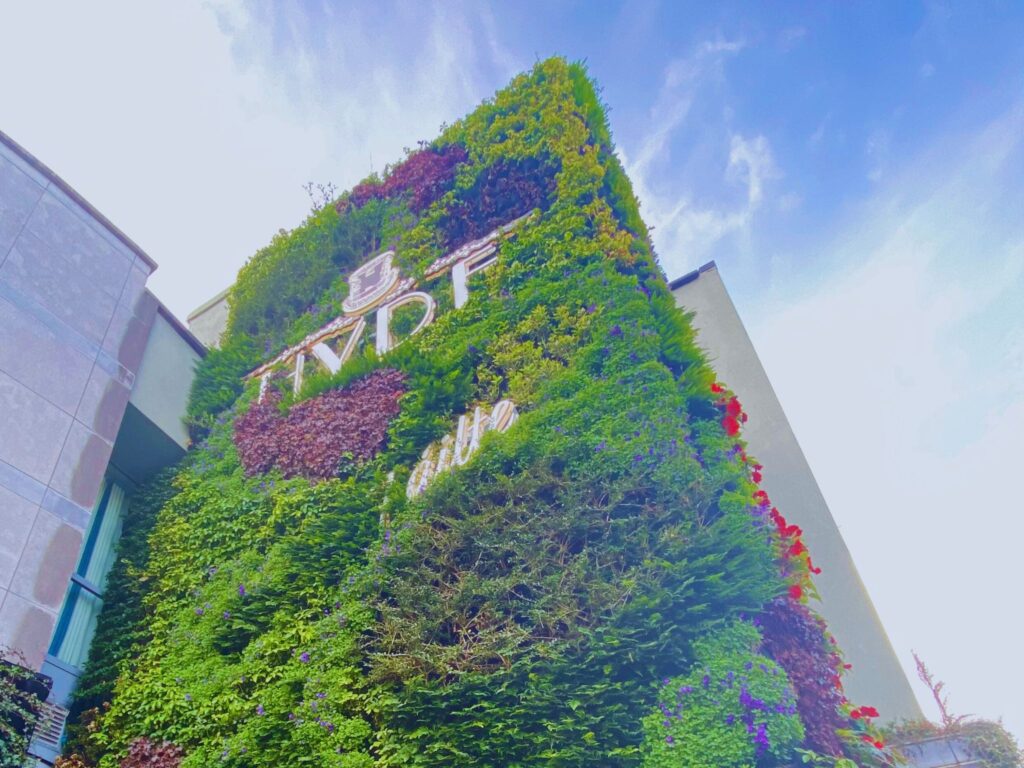 A tall building façade covered entirely in lush green and red foliage, forming a large living wall. The HYDE logo is prominently displayed among the plants near the top. The photo is taken from a low angle against a bright blue sky, highlighting the vertical garden’s vibrant colors and texture.
