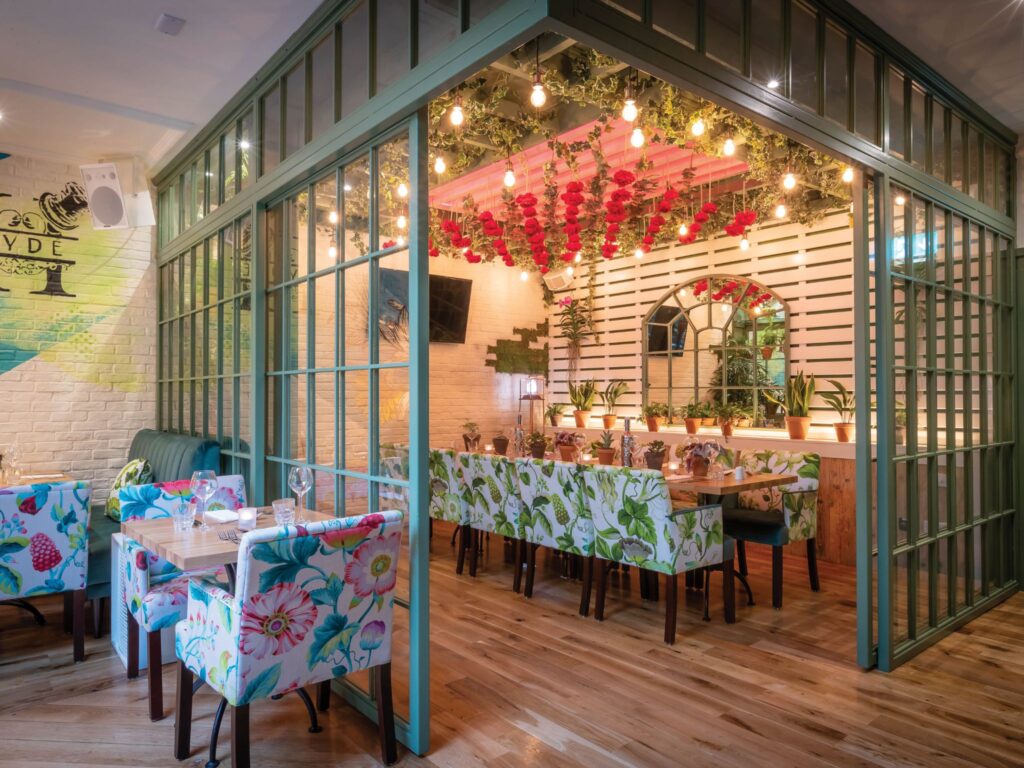 A vibrant, floral-themed restaurant interior featuring patterned chairs, long wooden tables, and green-framed glass partitions. The ceiling is decorated with hanging red roses and warm string lights, while potted plants line a shelf beneath an arched mirror on a white slatted wall. The space has a bright, botanical atmosphere with wooden flooring and colorful decor.