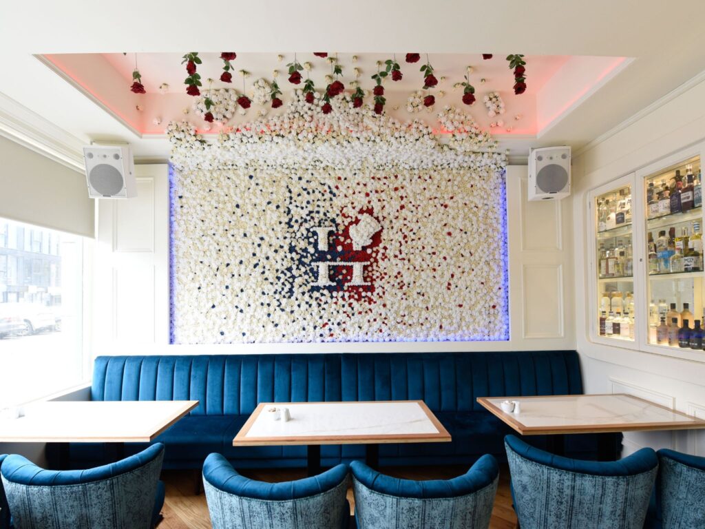 A bright, elegant bar interior featuring a long blue velvet banquette with marble-top tables and blue upholstered chairs. The focal point is a large wall covered in white, red, and blue flowers forming a decorative pattern with a central emblem. Hanging roses adorn the ceiling, and shelves of spirits are displayed in a glass cabinet on the right.
