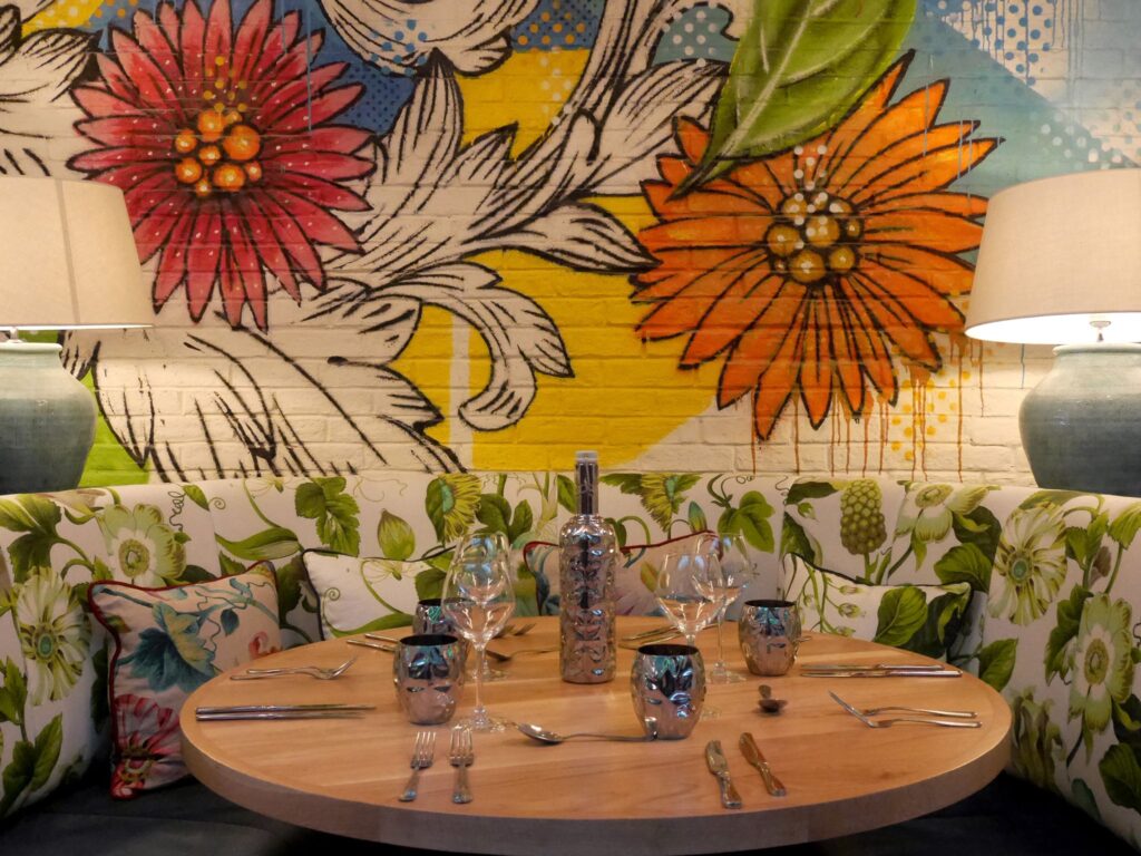 Vibrant restaurant table setting with floral-patterned seating, colorful flower mural on a brick wall, two large lamps on either side, and a round wooden table arranged with wine glasses, cutlery, and a decorative bottle at the center.