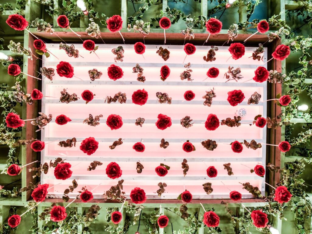 Ceiling installation featuring rows of bright red artificial roses and small clusters of foliage suspended against a softly glowing pink backlit panel, framed by greenery and exposed wooden beams.