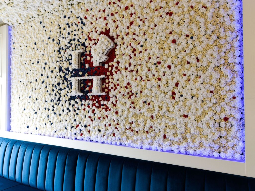 Large decorative wall covered in hundreds of white, red, and navy artificial roses arranged to form the Hyde logo, with soft purple backlighting and a deep blue velvet bench beneath it.