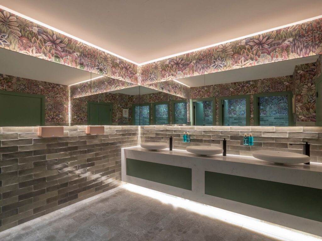 A stylish restroom featuring a long counter with four round vessel sinks and black faucets. The walls are decorated with botanical-patterned wallpaper and glossy greenish-brown tiles. Large mirrors stretch across the space, reflecting the vibrant design. Soft under-counter and ceiling lighting create a warm, modern ambiance.