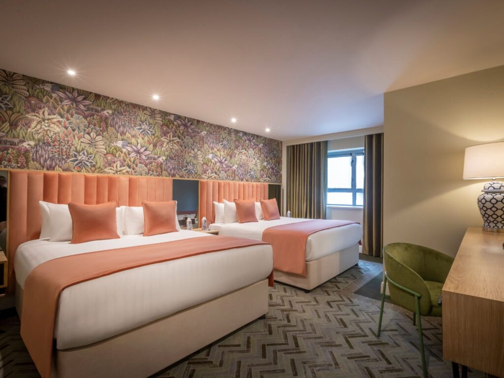 A spacious hotel room featuring two large beds with peach-colored throws and matching cushions, set against a vibrant floral accent wall. The room includes a patterned carpet, a wooden desk with a lamp, a green upholstered chair, and a window with curtains allowing natural light in.