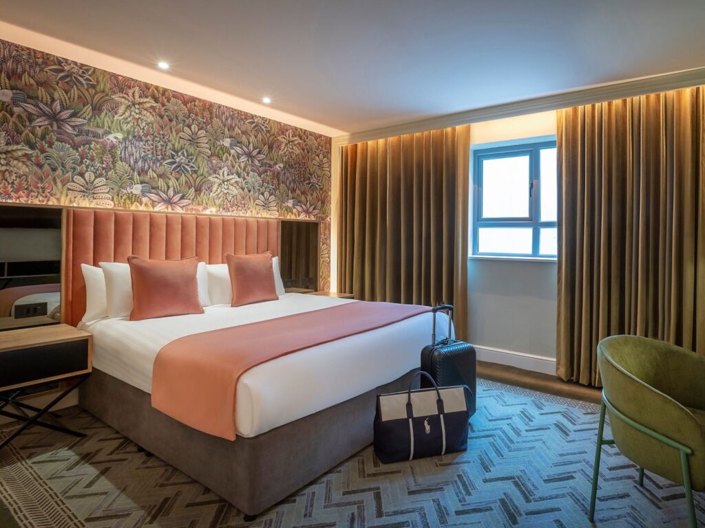 A stylish hotel room featuring a large bed with a peach-colored headboard, matching throw, and pillows. A bold botanical-patterned accent wall runs behind the bed. Floor-to-ceiling golden curtains frame a single window, and travel bags are placed neatly beside the bed. The room includes a green upholstered chair and patterned carpet, creating a warm and contemporary atmosphere.