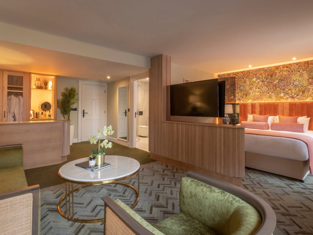 A spacious, warmly lit hotel suite featuring a seating area with green upholstered chairs and a round marble coffee table in the foreground. A wood-paneled partition holds a large flat-screen TV and separates the lounge from the bedroom, which includes a king-size bed with peach and white bedding and a floral accent wall. Built-in cabinetry, soft carpet, decorative lighting, and a glimpse of a modern bathroom contribute to the suite’s elegant, contemporary design.