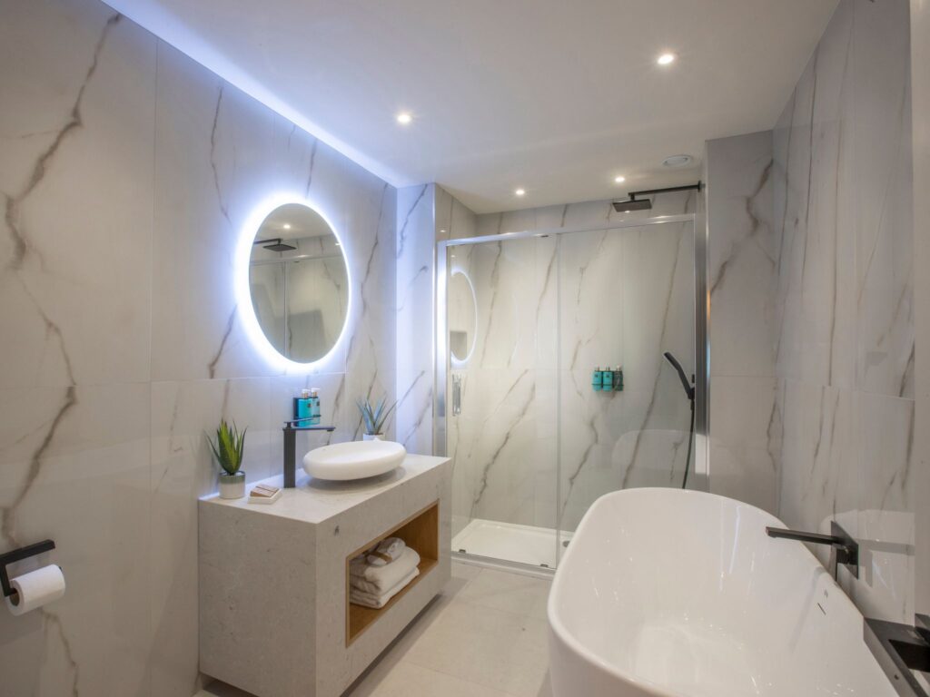 A modern, bright bathroom featuring large marble-patterned tiles, a freestanding white bathtub with a black faucet, and a spacious glass-enclosed walk-in shower. A circular backlit mirror hangs above a minimalist stone vanity with a vessel sink, toiletries, and small potted plants. Soft recessed lighting illuminates the clean, contemporary space.