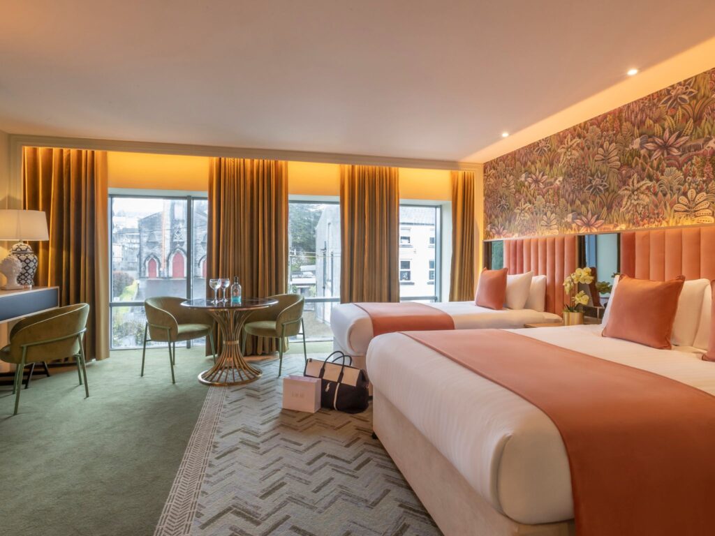 A spacious, warmly lit hotel room featuring two large beds with peach-colored throws and matching cushions. The headboard wall is decorated with vibrant botanical wallpaper. Floor-to-ceiling windows with golden curtains overlook nearby buildings. A round table with glasses and two green upholstered chairs sits near the windows, and additional seating and a lamp are placed along the left wall. Shopping bags rest on the floor between the beds.