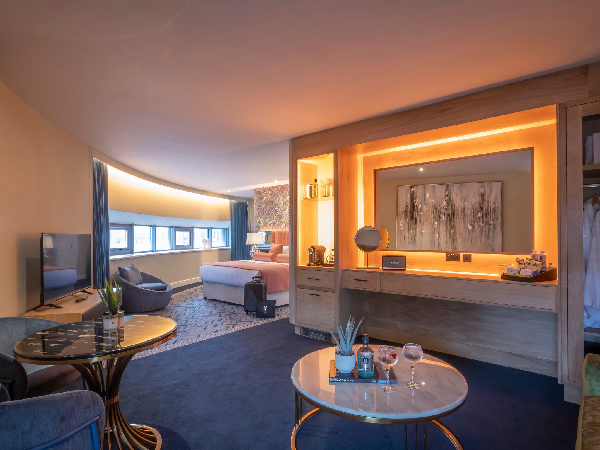 A modern, spacious hotel suite featuring a lounge area with round tables, upholstered chairs, and decorative plants. A built-in wooden bar and vanity area with warm backlighting and artwork sits to the right. In the background, a large bed with peach-colored bedding is partially visible, alongside a curved wall lined with small windows and long curtains. A TV stands on a low console to the left, and the room is illuminated with soft, ambient lighting.