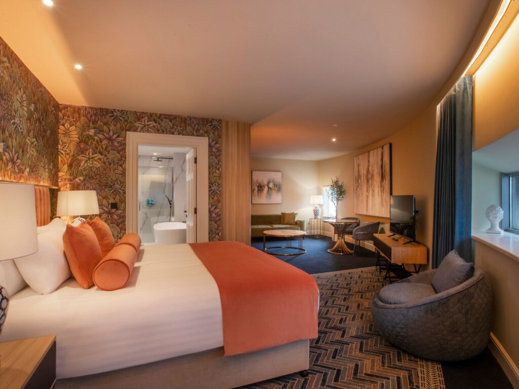 A spacious, warmly lit hotel suite featuring a large bed with peach and white bedding against a vibrant botanical accent wall. The room includes a seating area with a sofa and round tables, modern artwork on the walls, a TV on a wooden console, and a cozy cushioned chair near a small window. An open doorway reveals a bright, modern bathroom with a walk-in shower.