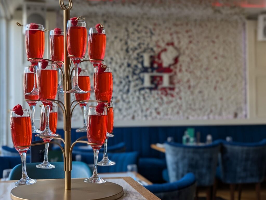 A gold stand holds multiple glasses of vibrant red cocktails, each garnished with a raspberry. The background features a blurred restaurant interior with blue seating and a textured white wall, creating an elegant, festive atmosphere.