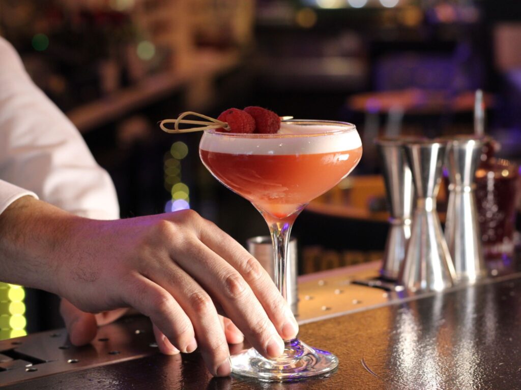 A bartender's hand places a vibrant, red cocktail garnished with raspberries on a bar counter. The dimly-lit background adds a cozy, elegant atmosphere.