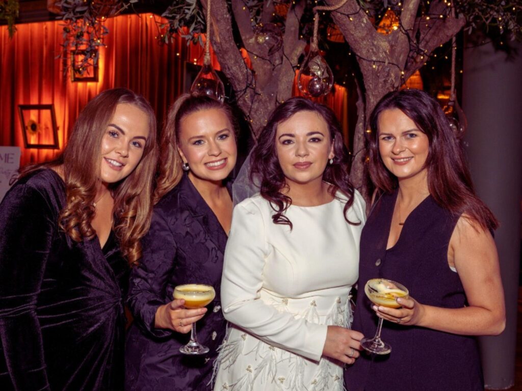 Four women in elegant dresses stand together, smiling and holding cocktails. The background features warm lighting and decorated trees, creating a festive atmosphere.