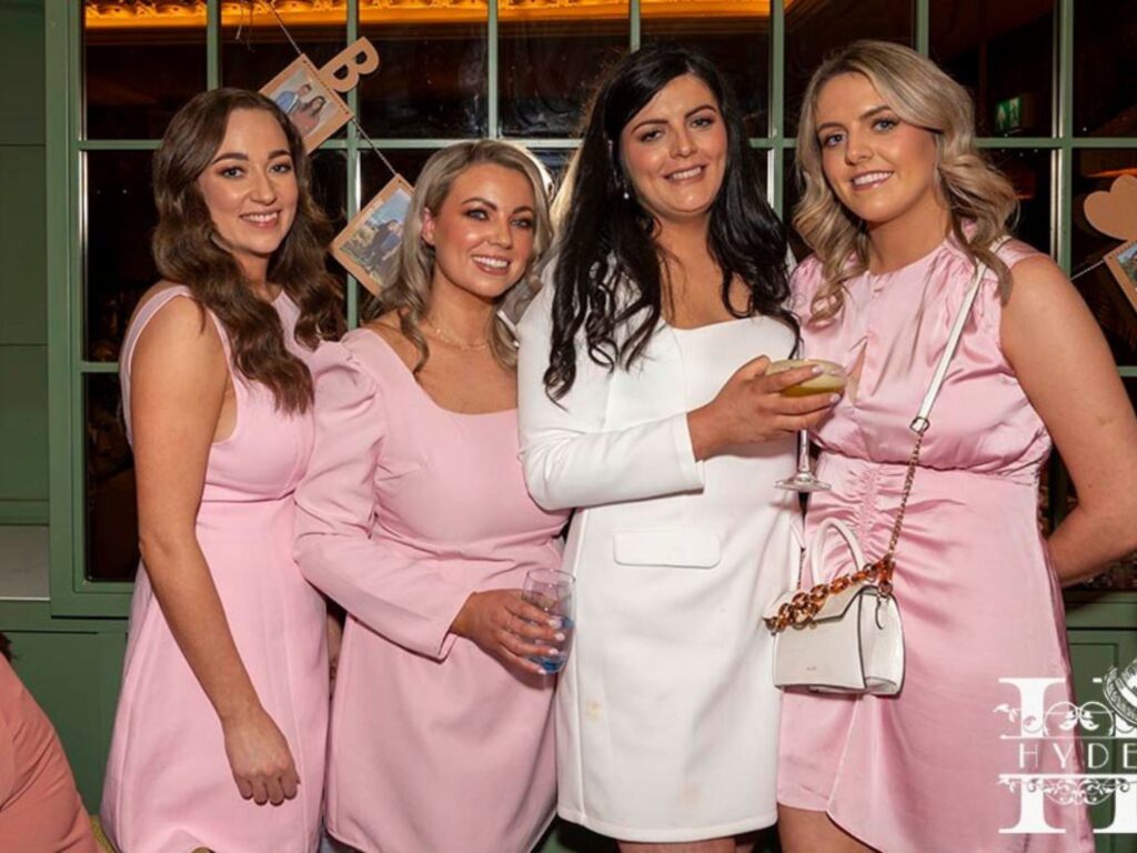 Four women pose together in a festive setting. Three wear light pink dresses, and one in white holds a drink. They smile warmly at the camera.