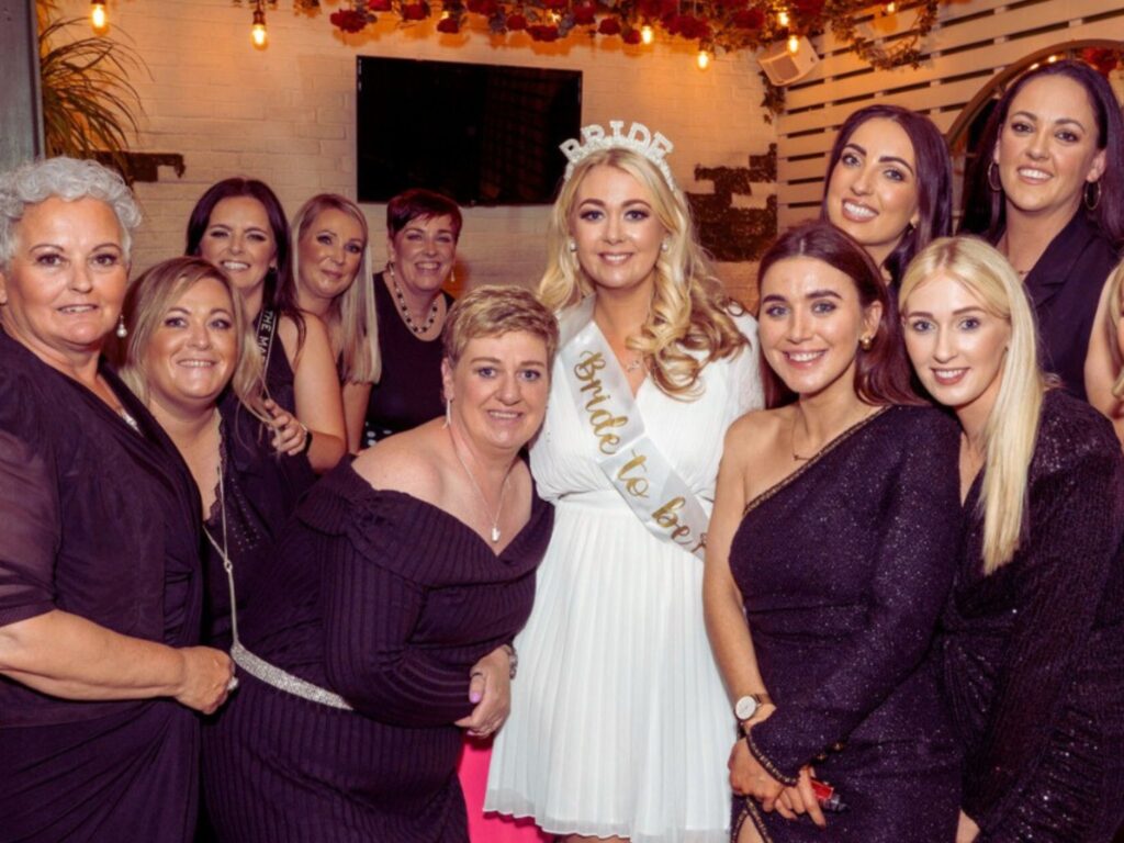 A group of women in black dresses surrounds a woman in a white dress and "Bride to be" sash. They are smiling, conveying a joyful celebration.