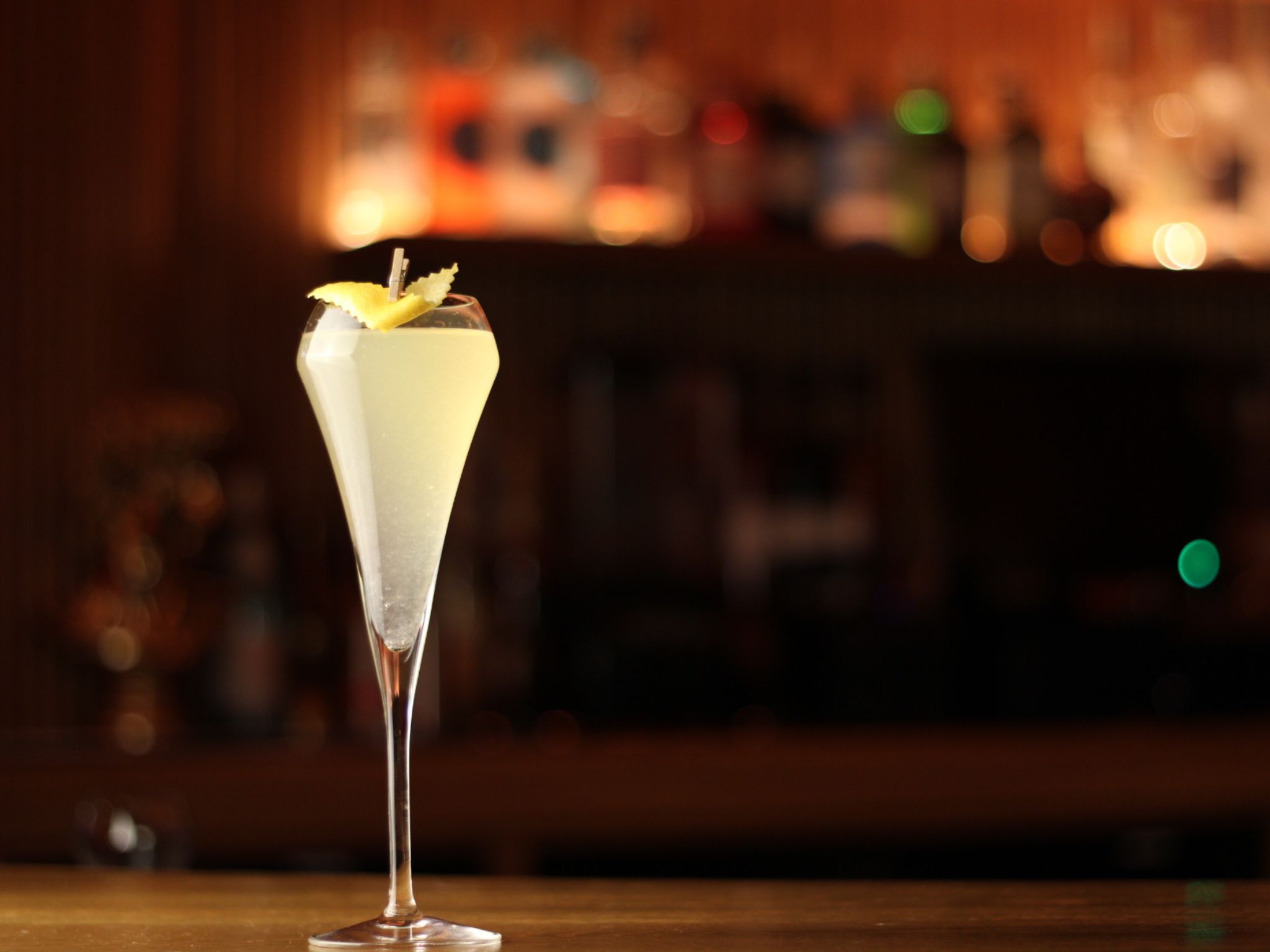 A tall, elegant flute glass with a pale yellow cocktail, garnished with a lemon twist, sits on a bar counter. The background is softly blurred and warmly lit.