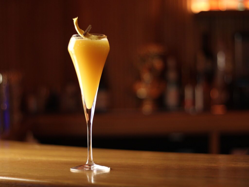 Elegant cocktail glass with a frothy, orange-yellow drink, garnished with a lemon twist, sits on a wooden bar, softly lit for a cozy ambiance.