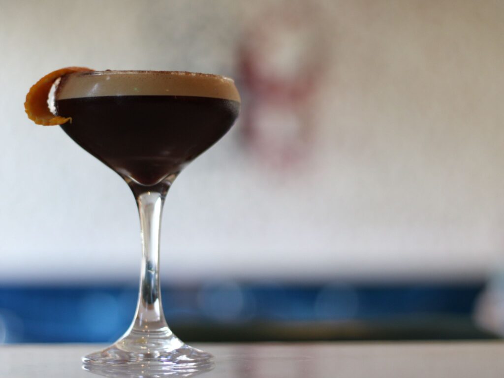A dark espresso martini with a foamy top sits in a sleek glass, garnished with an orange peel. The background is softly blurred, creating an elegant tone.