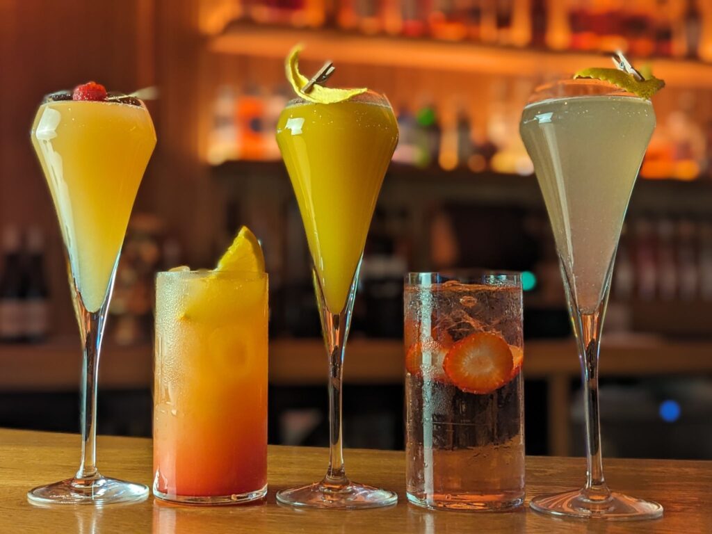 A variety of five colorful cocktails on a bar counter: three tall, slim glasses with garnishes and two short glasses with fruit slices, creating a vibrant and inviting mood.