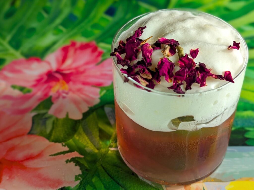 A glass with a cocktail with foam and dried rose petals atop, set against a vibrant floral background, conveying a refreshing, tropical vibe