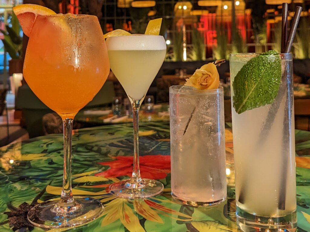 Four elegant cocktails on a vibrant floral table. An orange drink with grapefruit, a white drink with lemon, a clear drink with a flower, and a minty drink.