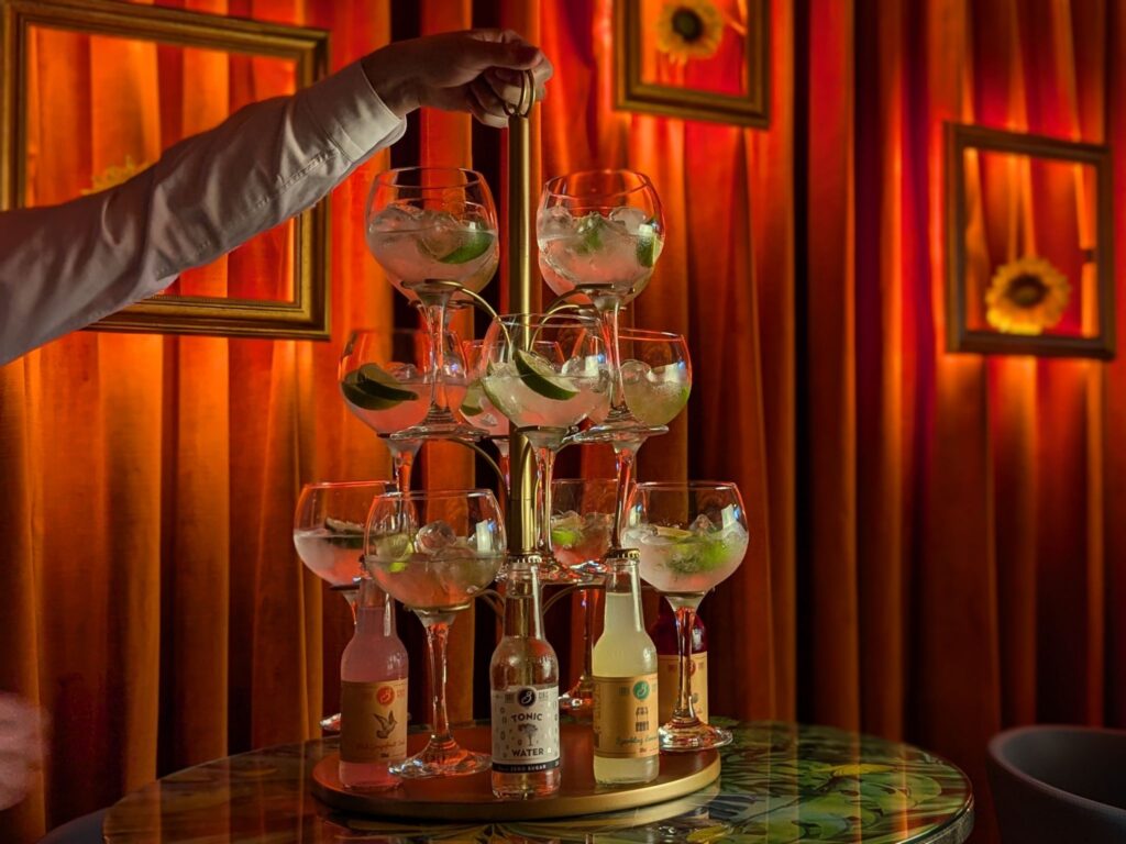 A person arranges gin glasses on a tiered stand, each garnished with lime and mint. Background features warm lighting and framed sunflowers.