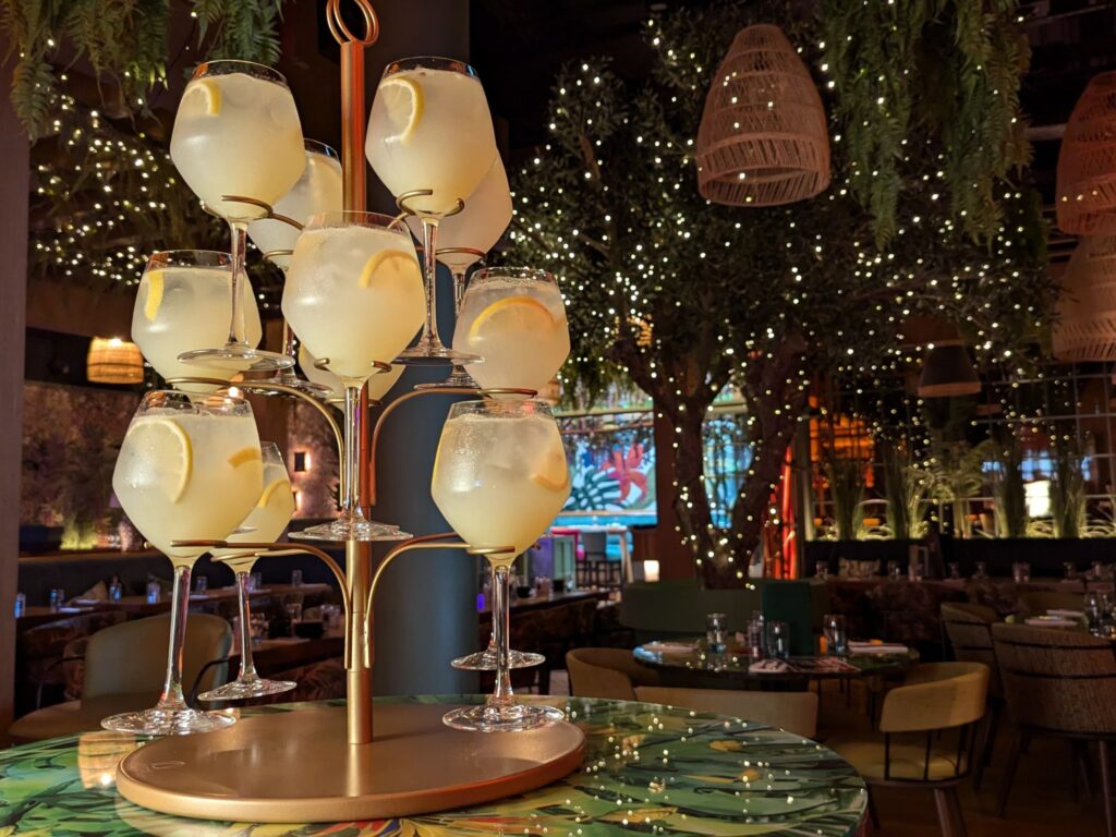 A tree-shaped stand holds glasses of lemon cocktails on a colorful table. The dimly lit restaurant is adorned with hanging plants and twinkling lights, creating an inviting, festive atmosphere.