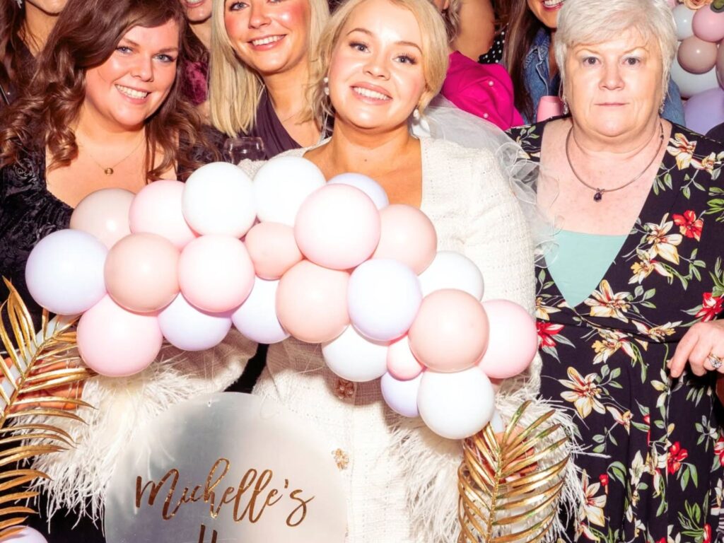 A group of smiling women pose with a pink and white balloon garland. They hold a sign reading "Michelle's Hen." The scene is joyful and celebratory.