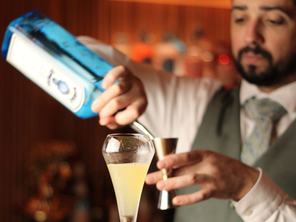 Bartender pouring blue gin into a measuring jigger over a yellow cocktail in a fancy glass. The background features a warmly lit bar ambiance.