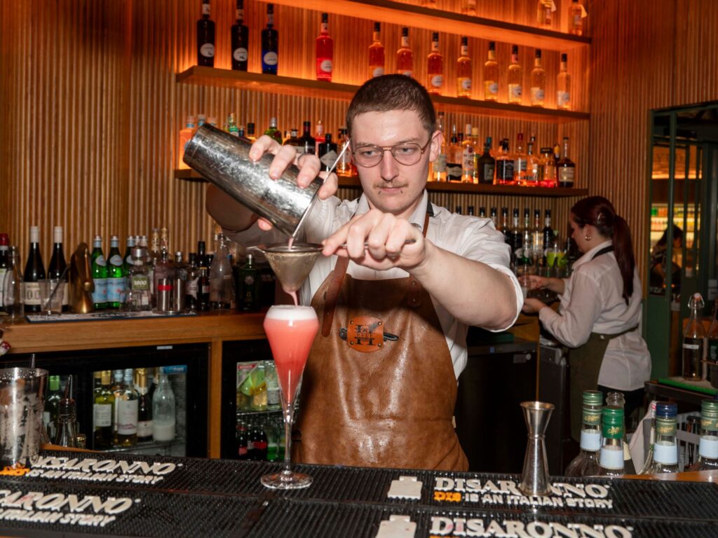 A bartender in a brown apron skillfully pours a pink cocktail through a strainer into a glass. Bottles line the bar shelves, creating a vibrant ambiance.