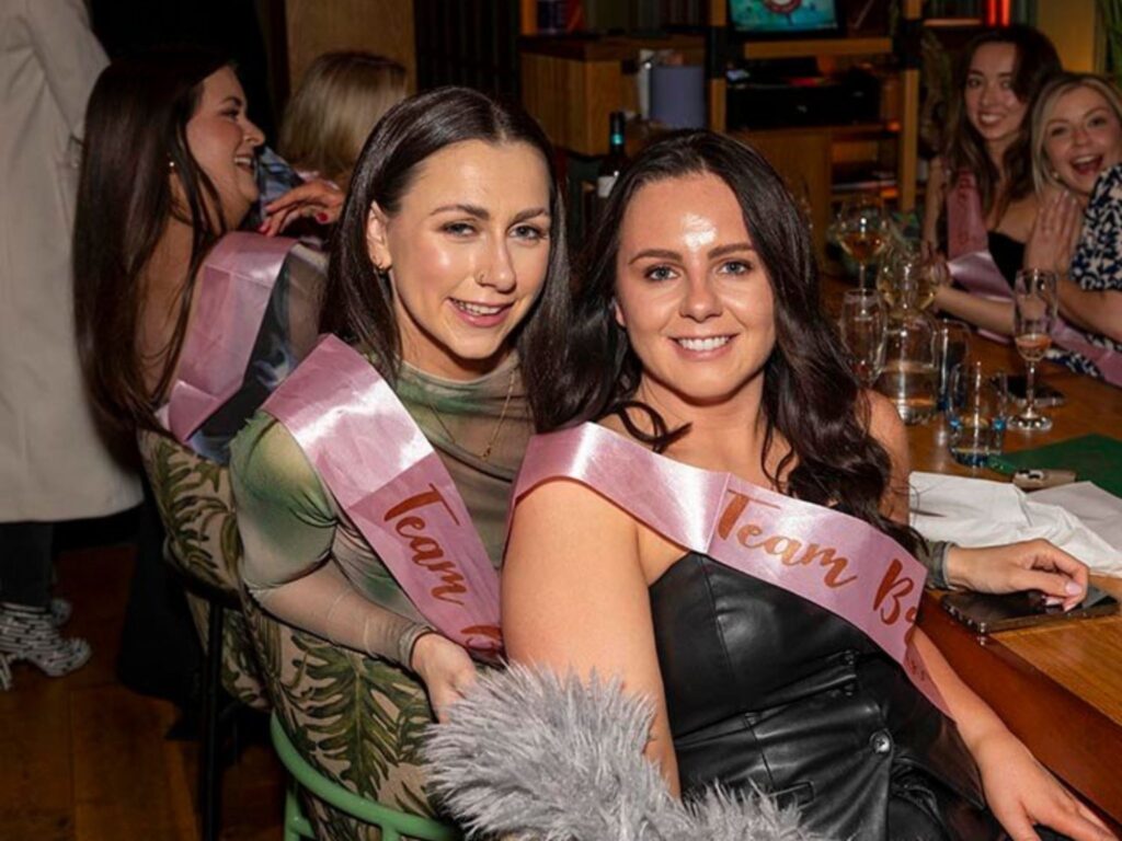 Two smiling women sit side by side at a table, wearing pink "Team Bride" sashes. The atmosphere is festive, with friends and drinks in the background.