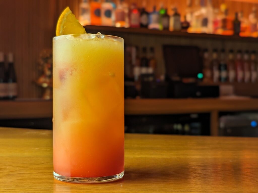 A vibrant tequila sunrise cocktail in a tall glass with an orange slice garnish sits on a wooden bar counter. The background shows a softly lit bar.