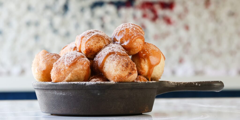 Skillet filled with golden-brown doughnut bites drizzled with caramel sauce and dusted with powdered sugar, served on a marble table.