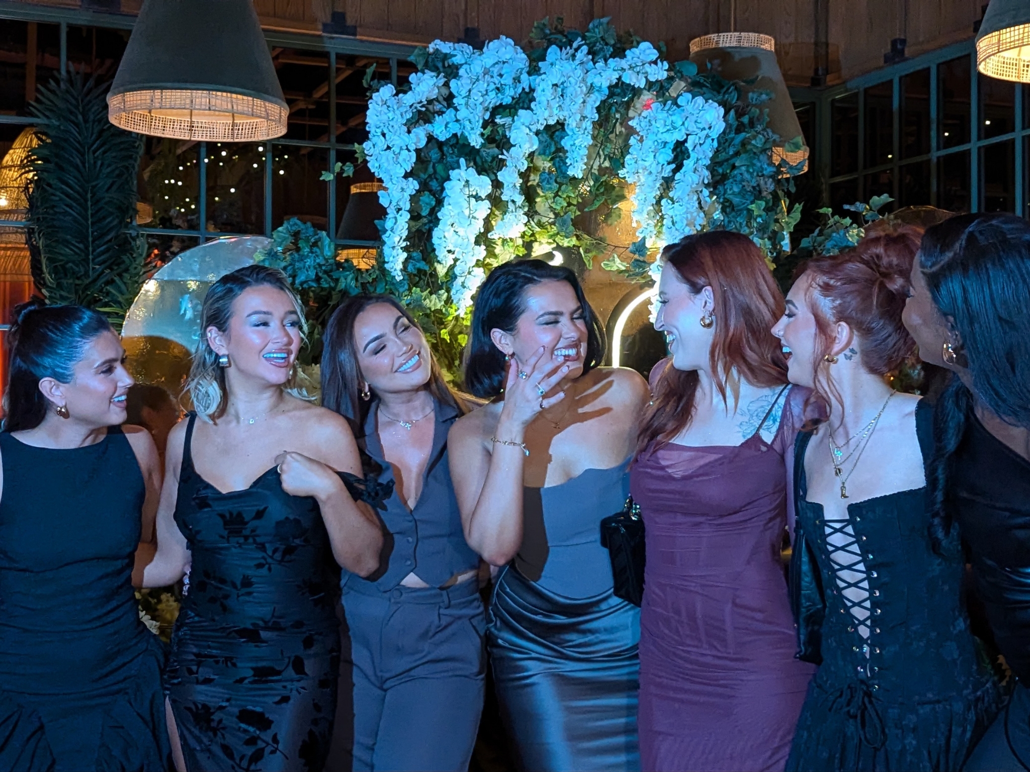 Group of women dressed elegantly in evening attire, laughing and posing together indoors against a lush floral backdrop with hanging lights and greenery.