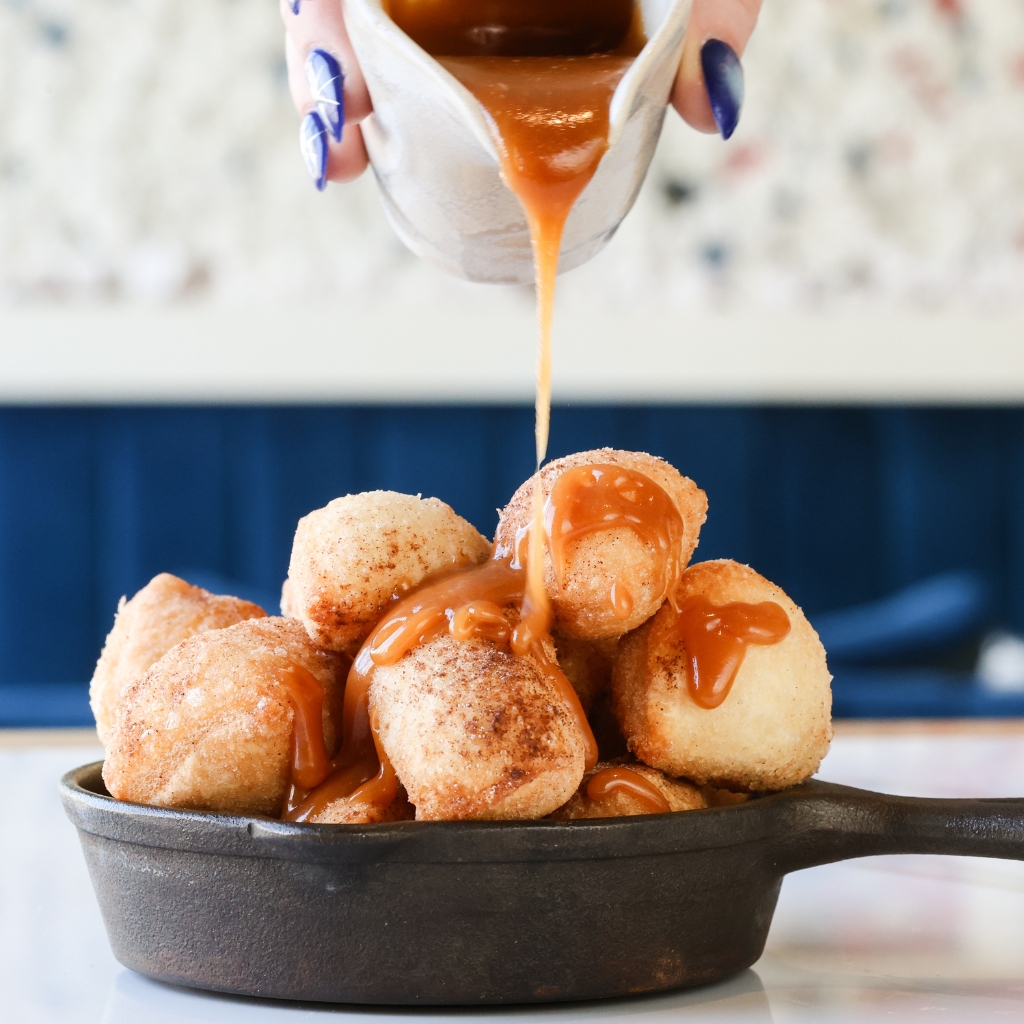 Close-up of caramel sauce being poured over a stack of sugar-dusted doughnut balls in a cast iron skillet, with a blurred background of a blue booth and light-colored wall.