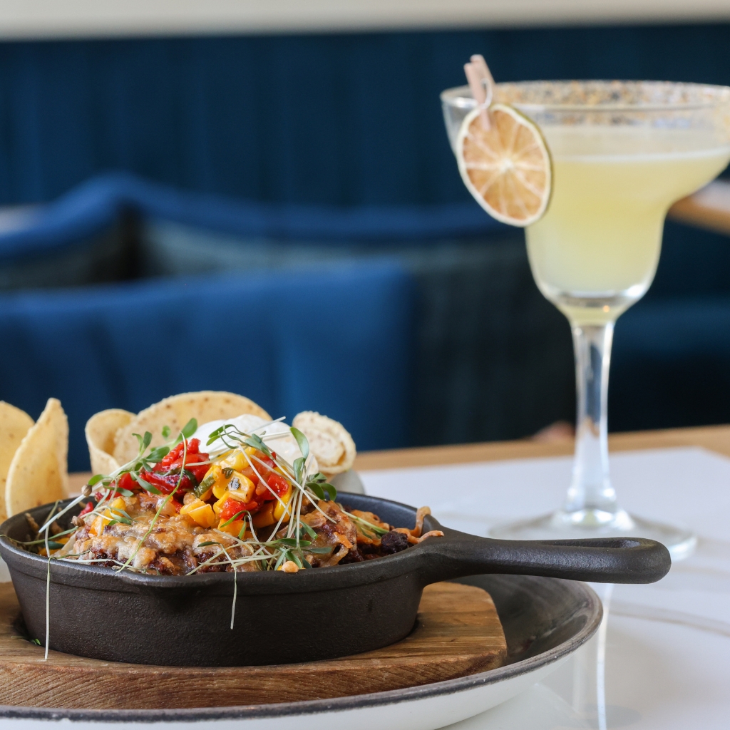 A skillet filled with a colorful dish topped with fresh herbs, corn, and red peppers, served with tortilla chips on the side. A margarita garnished with a dried lime slice sits beside it on a table with some blue upholstered chairs in the background.