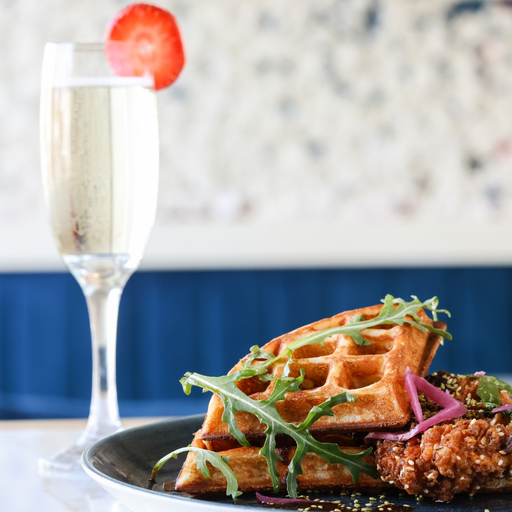 Close-up of a brunch dish featuring crispy fried chicken and golden waffles garnished with arugula and pickled onions, served on a dark plate beside a glass of champagne with a strawberry slice on the rim.