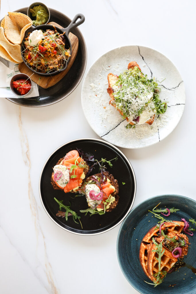Flat lay of four beautifully plated brunch dishes on a white marble table, including loaded nachos in a skillet, avocado toast topped with cheese, smoked salmon on brown bread, and chicken and waffles garnished with greens and pickled onions.