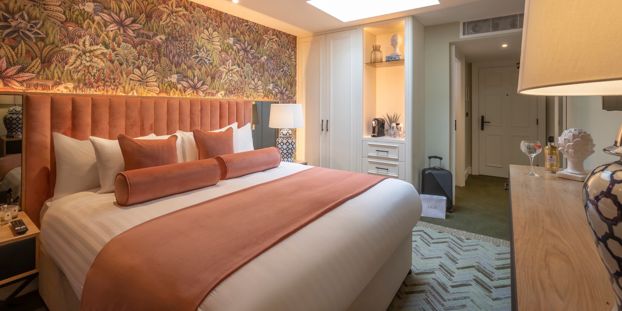 HYDE hotel room- Elegant hotel room featuring a large bed with peach-colored bedding and matching pillows, a floral-patterned accent wall, soft lighting, and built-in cabinets with a coffee station and decor.