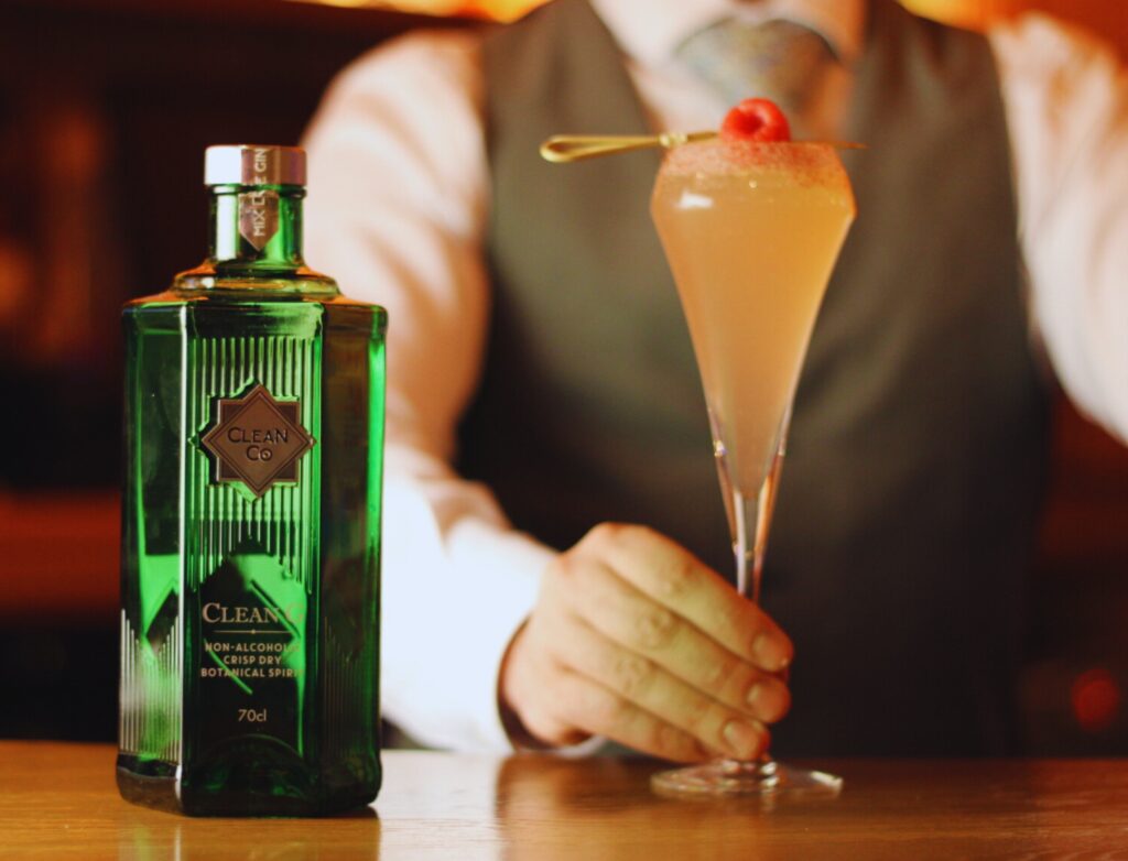 Bartender in a vest and tie presenting a pink cocktail garnished with a raspberry beside a green bottle labeled “Clean Co – Non-Alcoholic Crisp Dry Botanical Spirit” on the Jekyll bar counter.