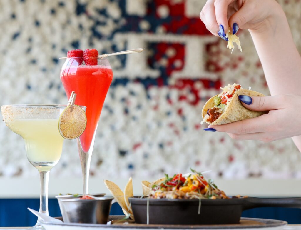 Close-up of a person adding cheese to a taco filled with guacamole and salsa, with two colorful cocktails—a red raspberry drink and a yellow margarita—beside a skillet of loaded nachos on a table.