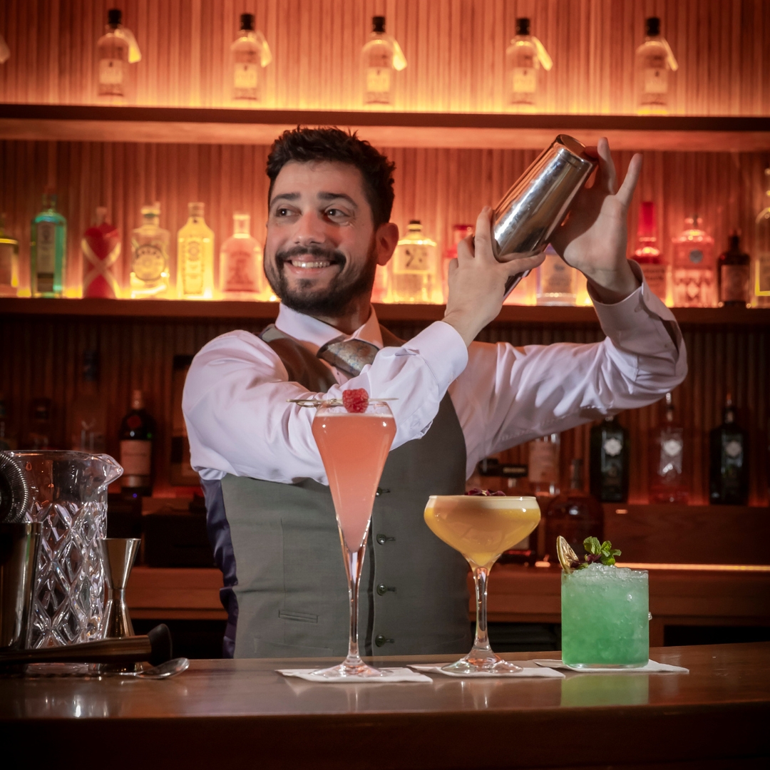 Smiling bartender in a vest and tie shaking a cocktail shaker behind the bar, with three colorful cocktails—pink, yellow, and green—lined up on the counter in front of illuminated liquor bottles.