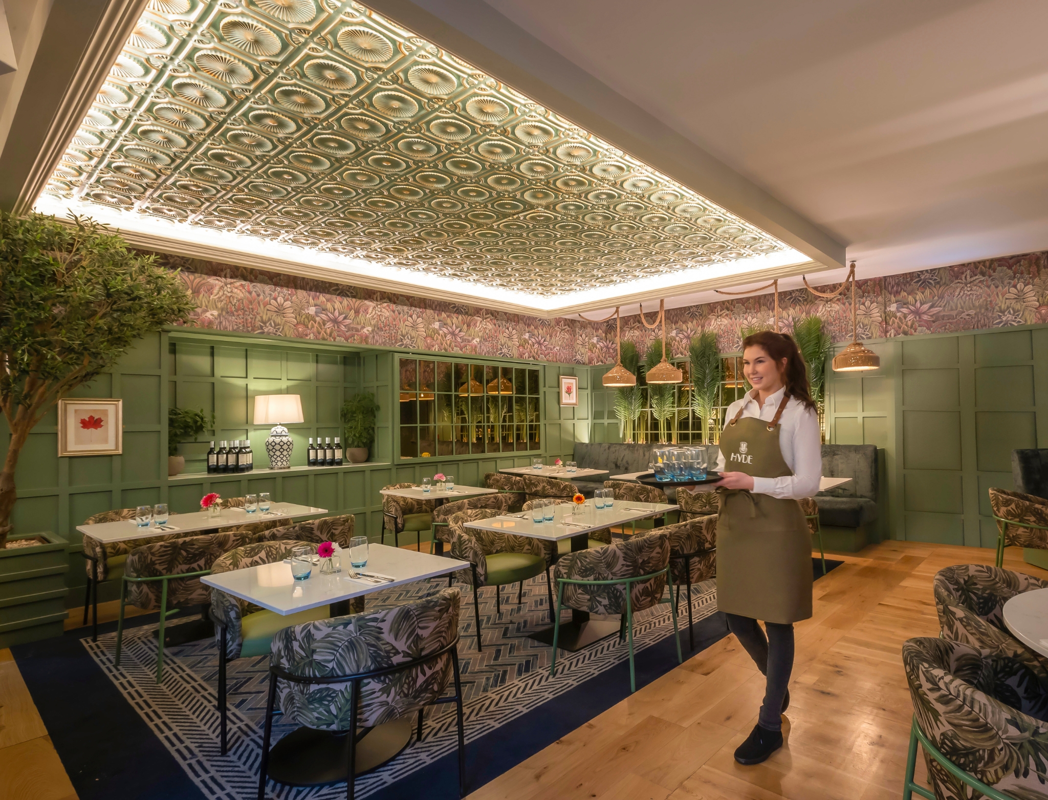 HYDE- Elegant restaurant interior with green paneled walls, floral wallpaper, patterned ceiling, and botanical-themed chairs. A smiling server wearing a green apron holds a tray of drinks, standing among neatly set tables decorated with small flowers and blue glassware.