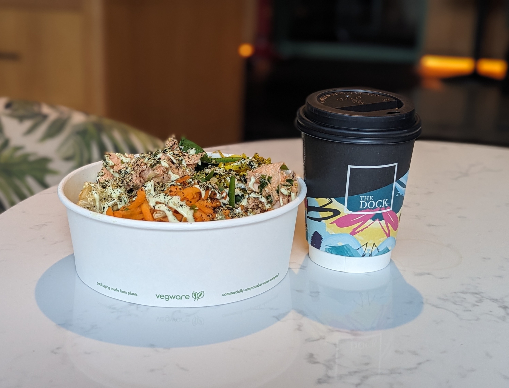 A takeaway meal in a white Vegware bowl from WYLDE, filled with mixed vegetables, grains, and sauce, placed next to a colorful coffee cup labeled “The Dock” on a marble table.